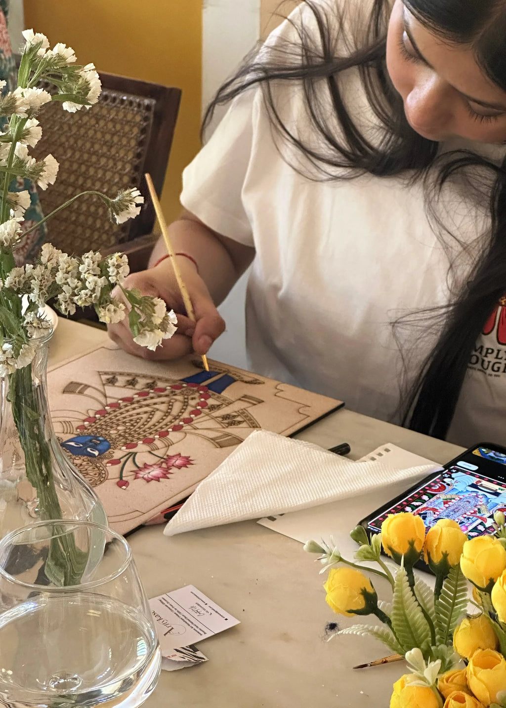 Woman painting a Lord Krishna Pichwai devotional art design at a table with flowers - Arttykase Indian folk art mindfulness kit for beginners