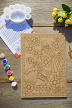 Arttykase Madhubani bird painting kit flat lay on a wooden surface - pre-etched A4 MDF board with two birds and floral folk art design, 12 acrylic paint pots, mixing palette, fine brush and Madhubani art instruction manual beside yellow flowers