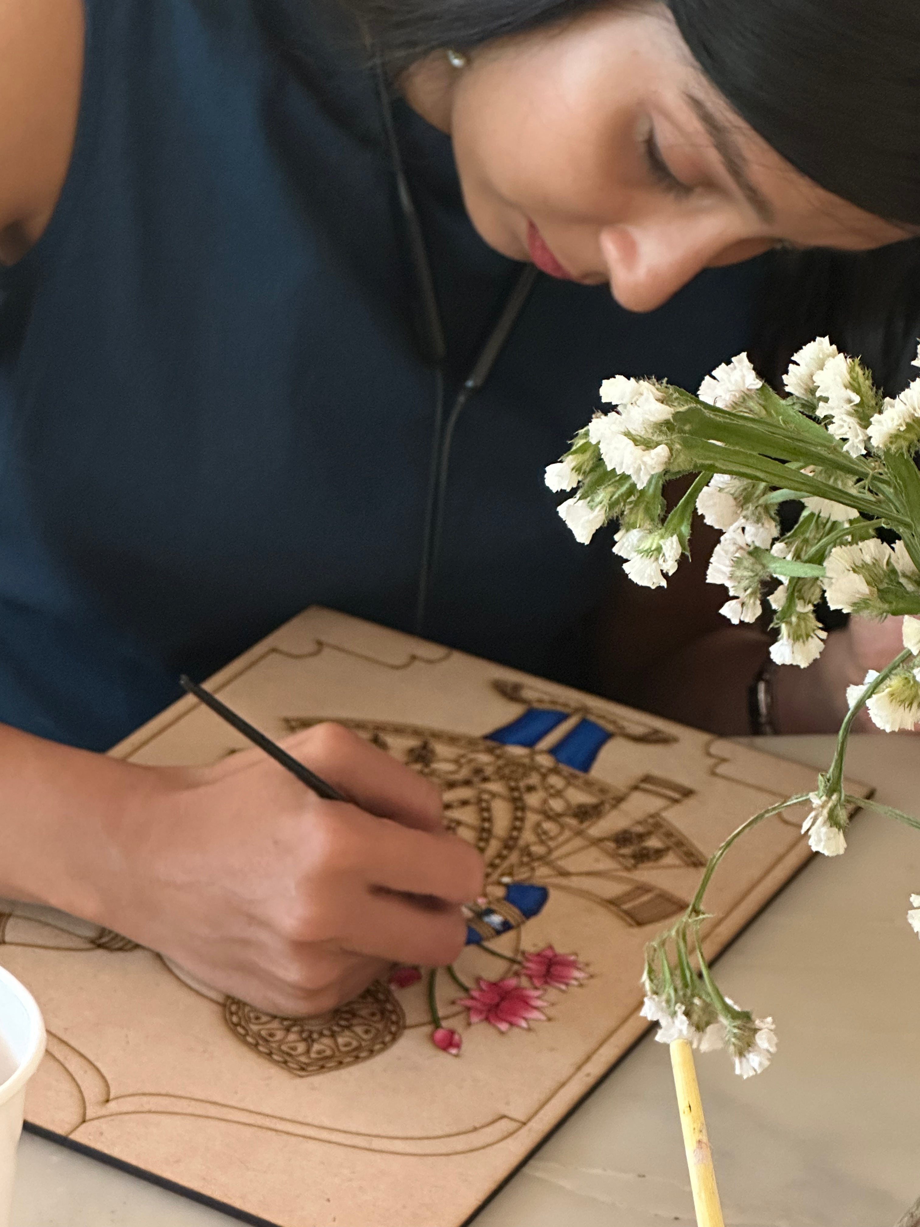 Woman painting an intricate Pichwai mandala design on a pre-marked MDF wooden board with a fine brush beside white flowers - Arttykase Indian folk art DIY kit lifestyle